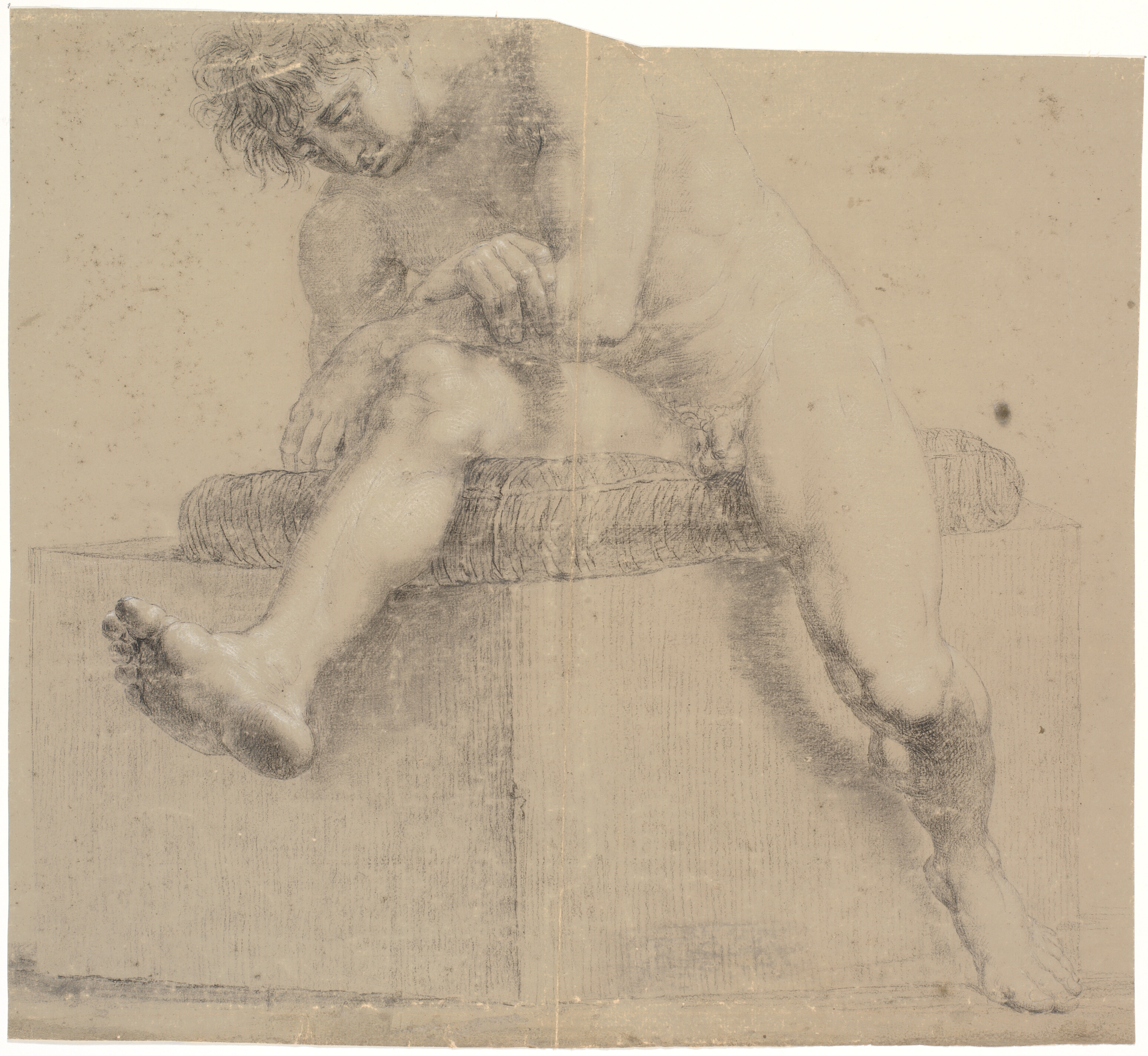 Siddende model på lav plint med pude. Armene krydset. Venstre ben bøjet, højre ben udstrakt